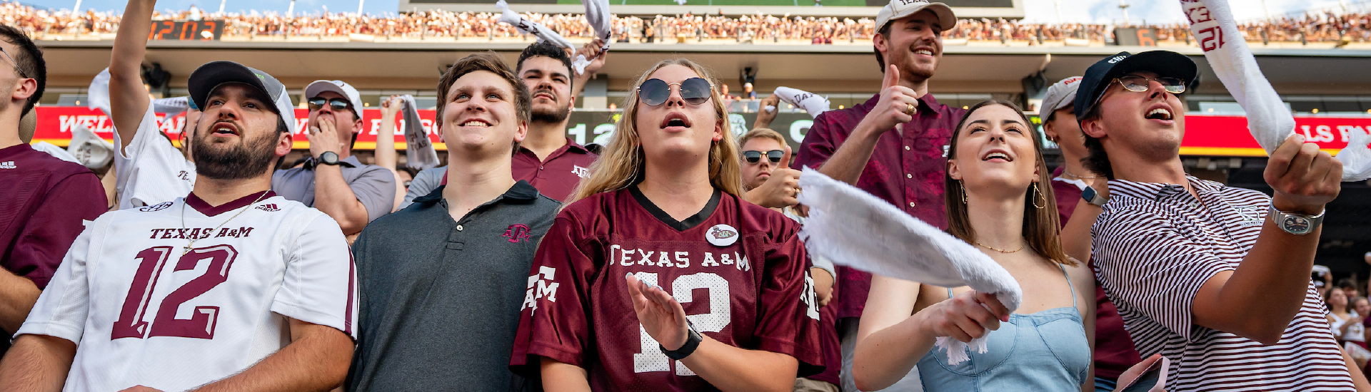 Students standing in the student section at an Aggie Football game at Kyle Field. Some are cheering and clapping, and others are waving 12th Man Towels.