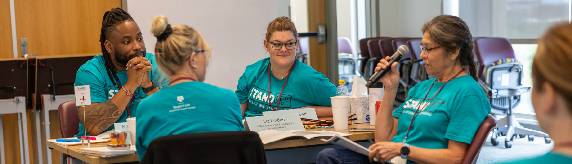 A staff member sitting at a table with others, speaking into a microphone during a Stand Up workshop