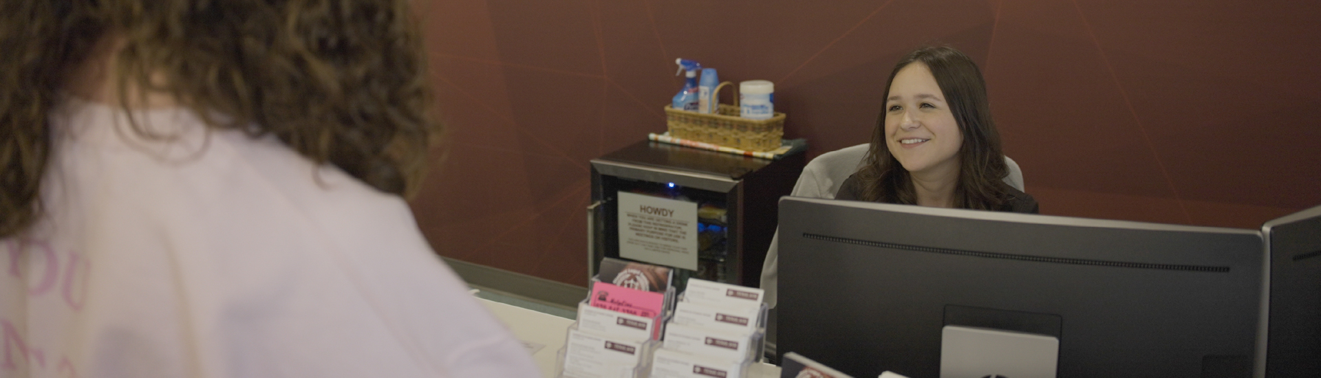 A Student Life student worker sitting behind a desk and computer, helping another student in need at the front desk.