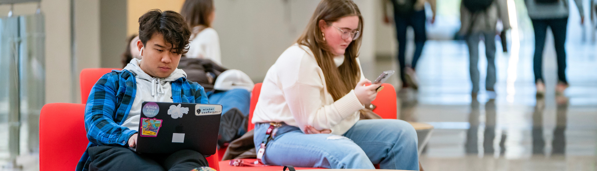 Two students sitting in Zachry, studying. One is on their laptop, the other is on her phone.