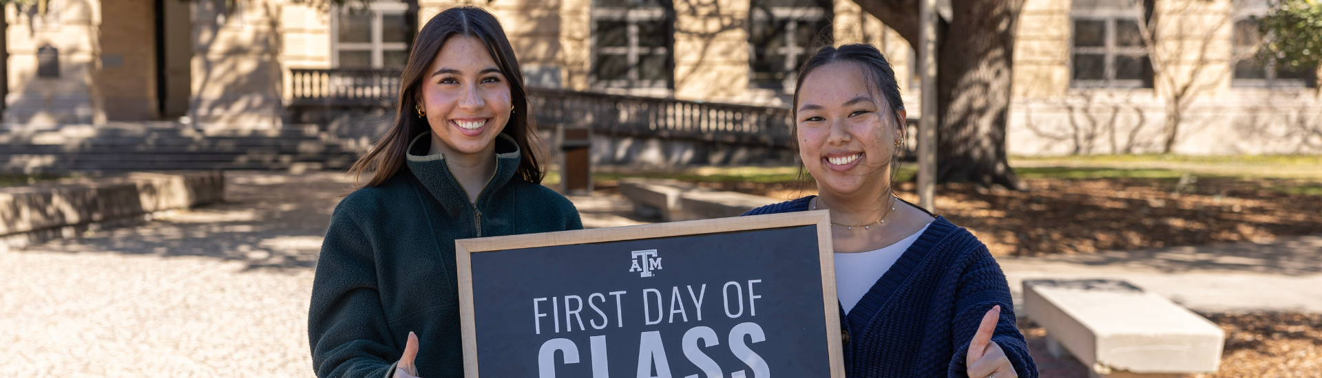 Two girls smiling and giving a "Gig 'em" thumbs up in front of the Academic Building holding a chalkboard that reads "First Day of Class Spring 2025."