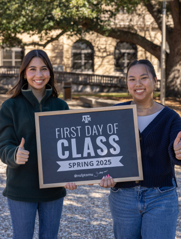 Two girls smiling and giving a "Gig 'em" thumbs up in front of the Academic Building holding a chalkboard that reads "First Day of Class Spring 2025."