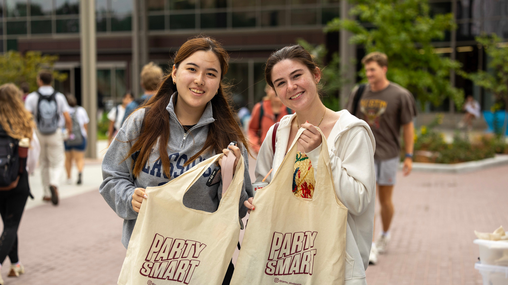 Two students holding canvas "Party Smart" tote bags outdoors on campus plaza with other students walking in background.