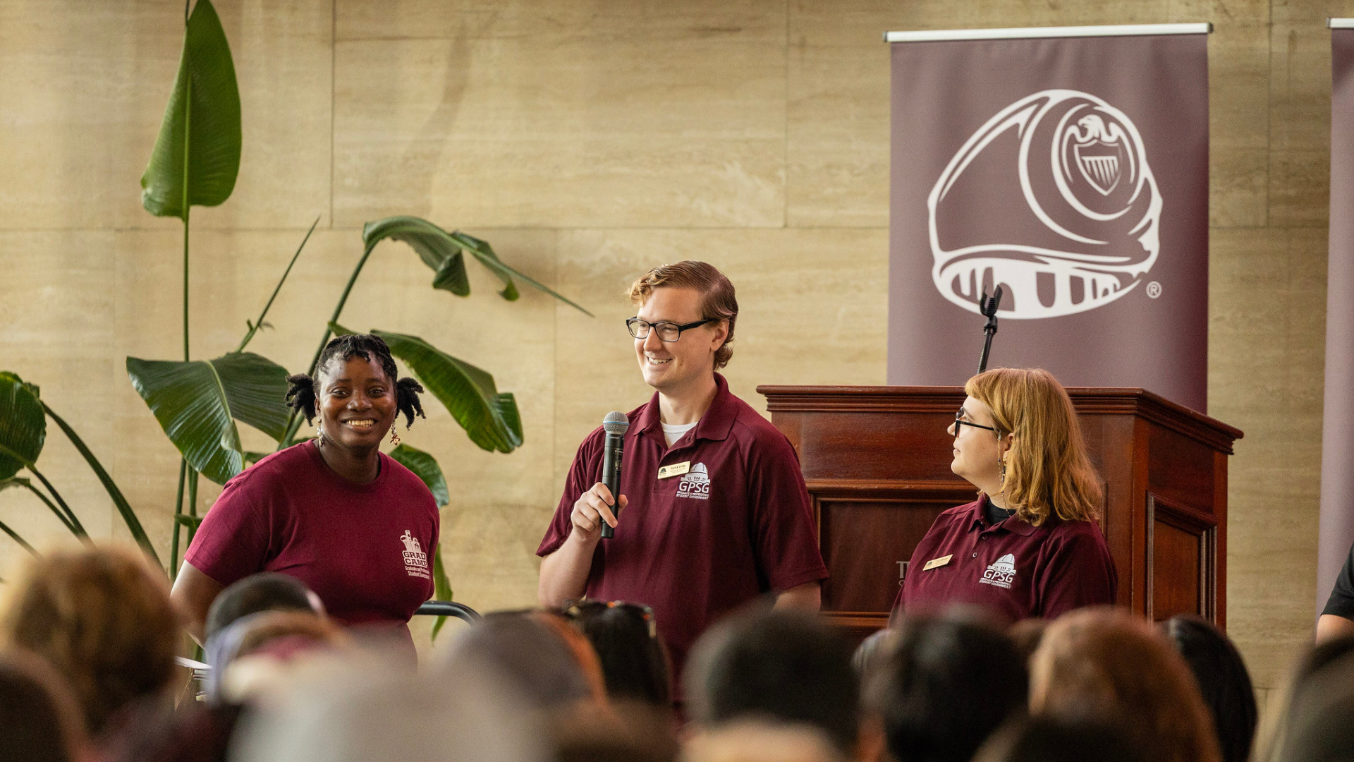 Three student leaders in maroon polos speaking to seated audience during Grad Camp with Texas A&M banner and in background.