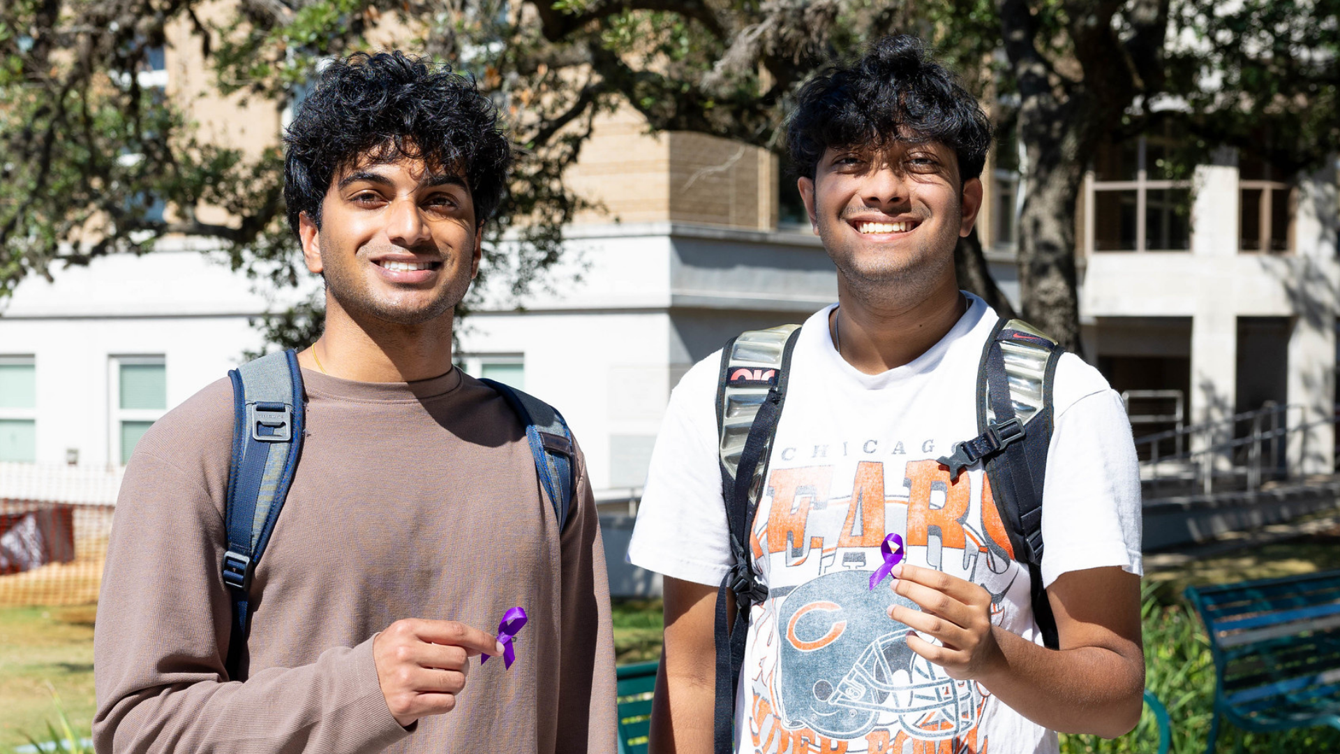Two students wearing backpacks and holding purple awareness ribbons posing together outdoors on campus with building visible behind them.
