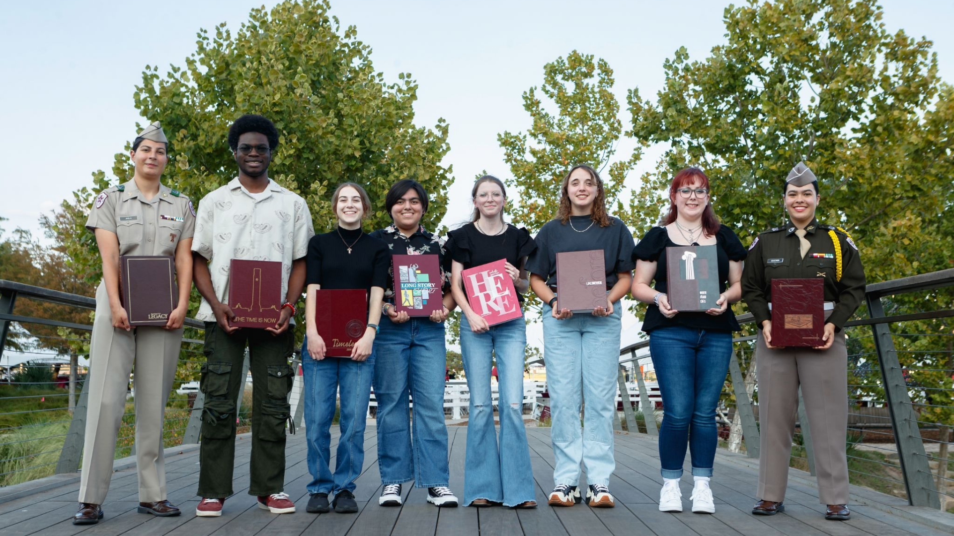 Eight students in standing in line on the bridge in Aggie Park holding maroon Aggieland Yearbooks with trees in background.