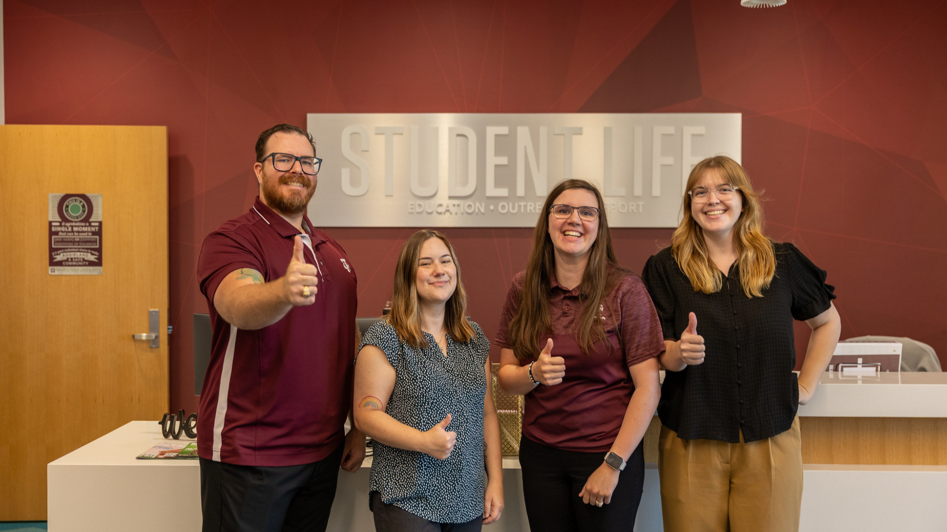 Four staff members giving thumbs up gesture indoors in front of maroon Student Life wall.