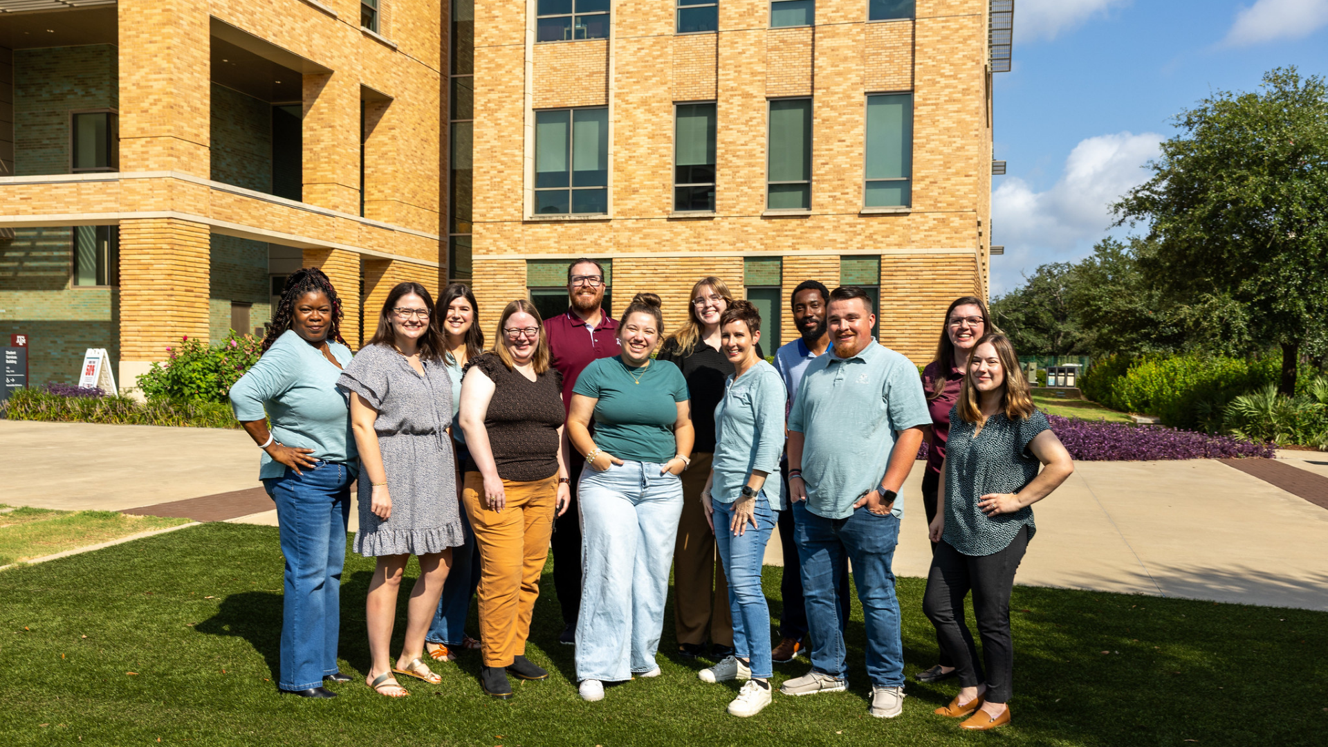 Staff team posing outdoors in front of Student Services Building with landscaping on sunny day.